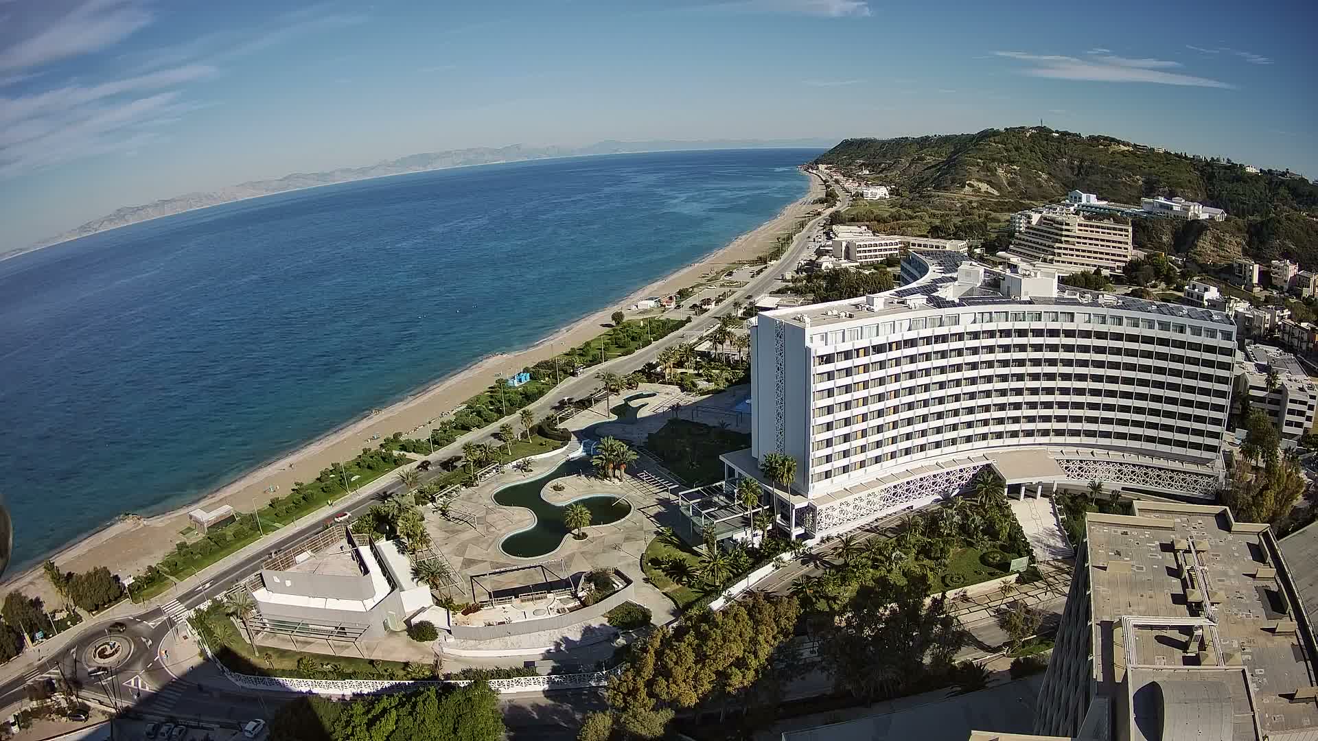 An aerial view captures a sunny day on a coastal resort town, featuring a long sandy beach, brilliant blue sea, and numerous white hotel buildings nestled along the shore and climbing verdant hills under a clear sky with scattered wispy clouds.