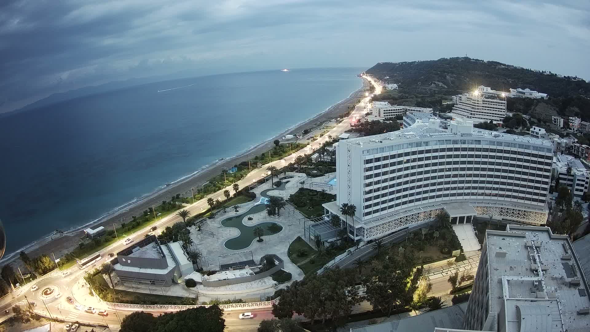 An aerial view captures a sunny day on a coastal resort town, featuring a long sandy beach, brilliant blue sea, and numerous white hotel buildings nestled along the shore and climbing verdant hills under a clear sky with scattered wispy clouds.