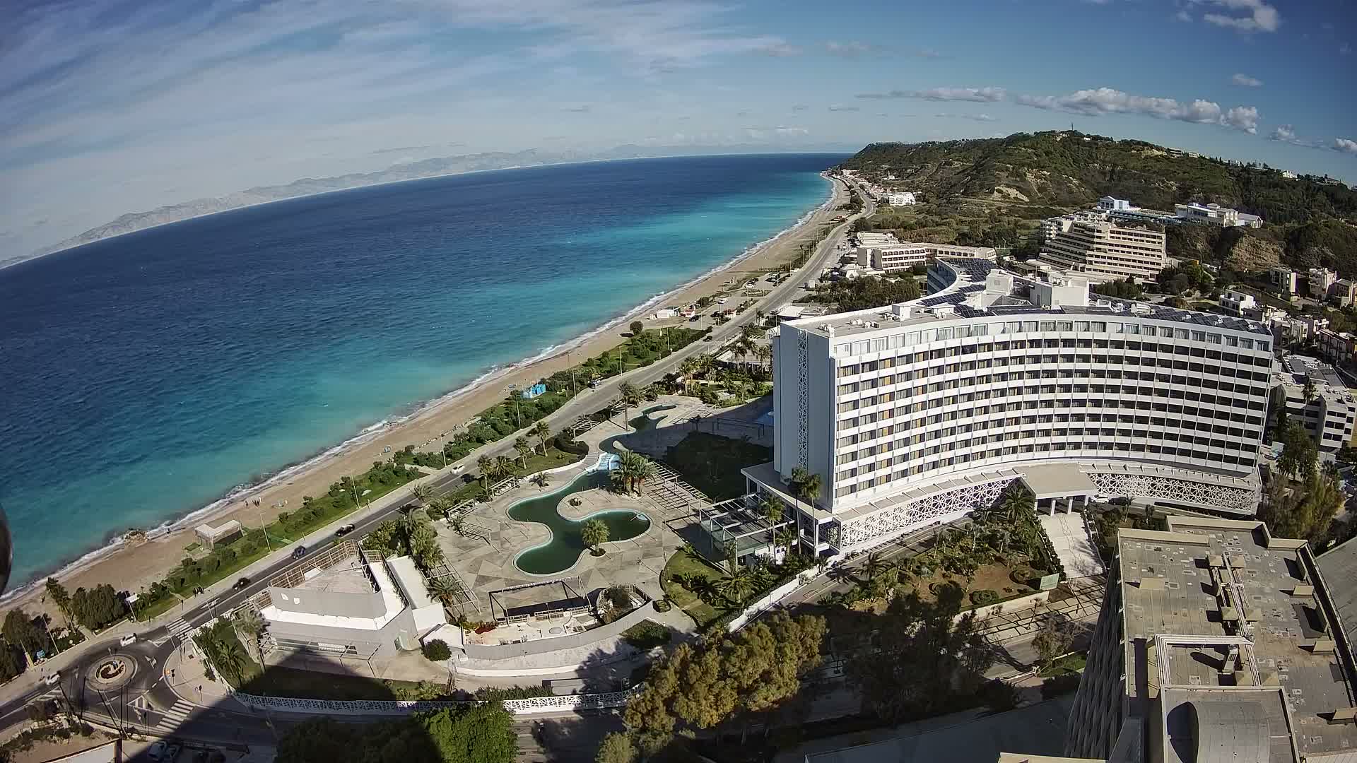 Under a clear blue sky with scattered white clouds, an aerial view captures a sunny coastal landscape featuring a sandy beach, clear turquoise ocean, a prominent white hotel complex with pools and gardens, and a coastal road winding along the shore.