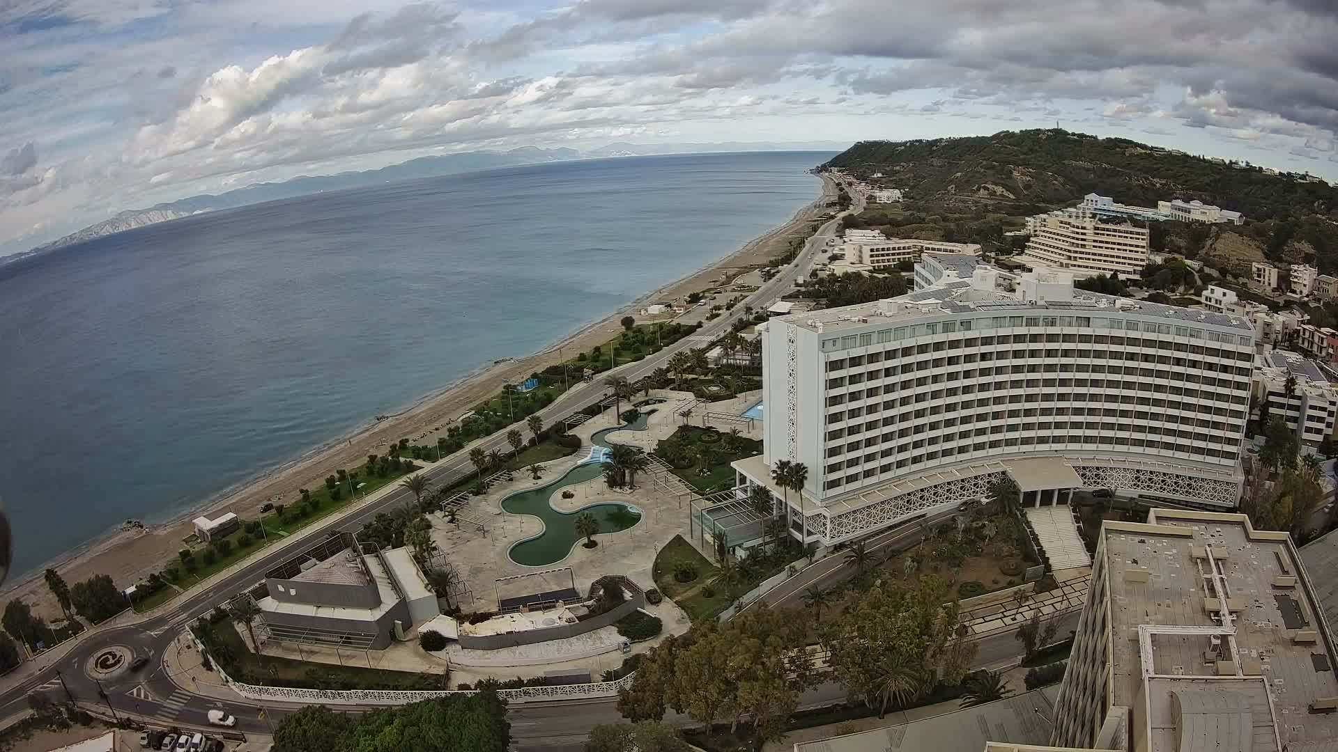 Under a clear blue sky with scattered white clouds, an aerial view captures a sunny coastal landscape featuring a sandy beach, clear turquoise ocean, a prominent white hotel complex with pools and gardens, and a coastal road winding along the shore.
