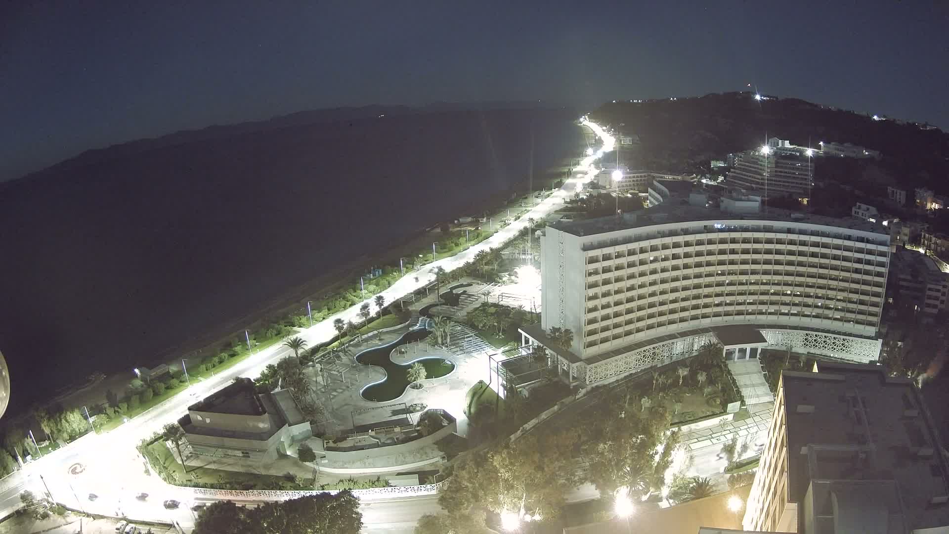 An aerial night view reveals a brightly lit coastal resort with a large hotel, intricate swimming pools, and a lamp-lined road running alongside the dark sea, all set against illuminated hills under a clear sky.