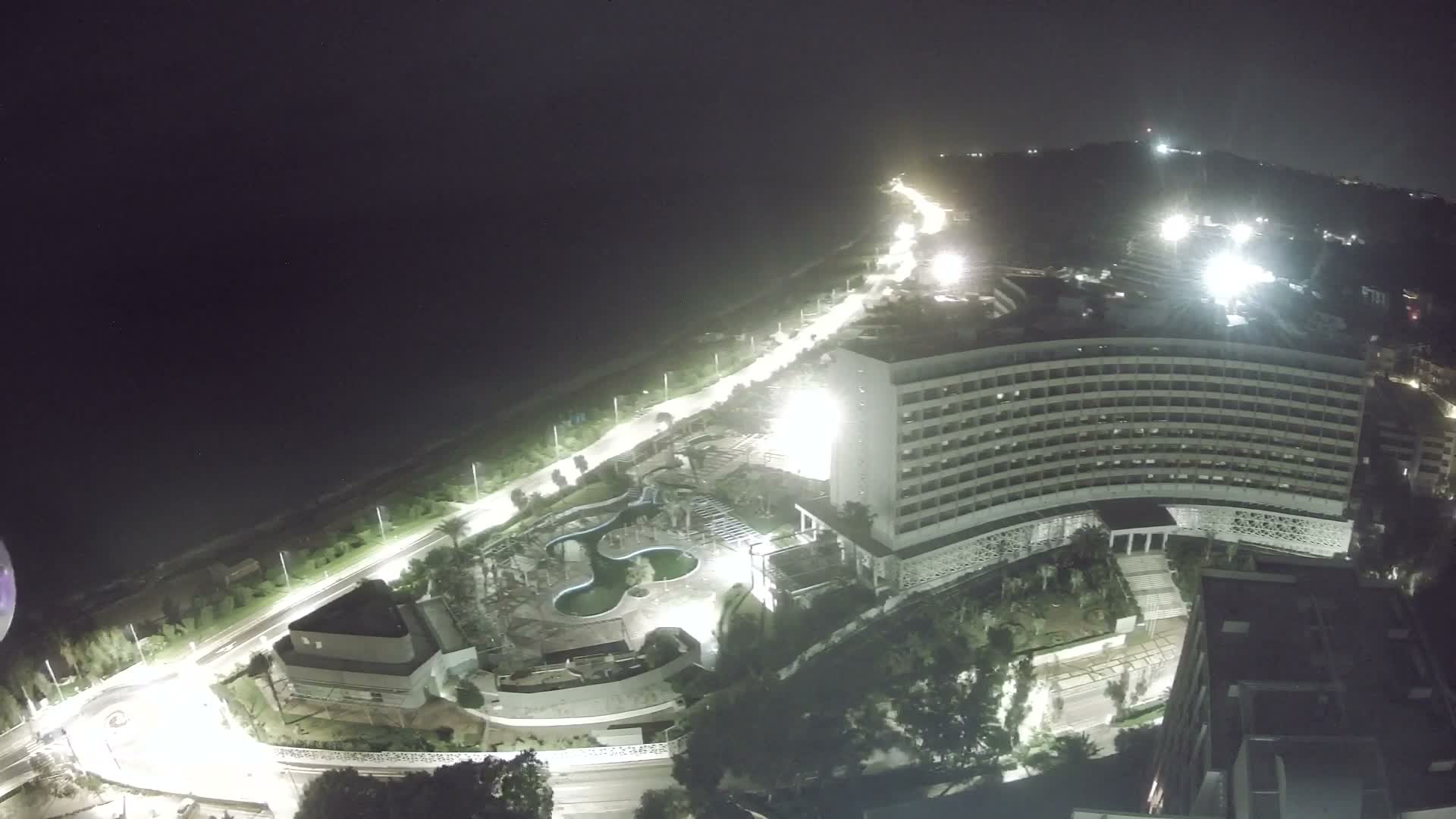 An aerial night view reveals a brightly lit coastal resort with a large hotel, intricate swimming pools, and a lamp-lined road running alongside the dark sea, all set against illuminated hills under a clear sky.