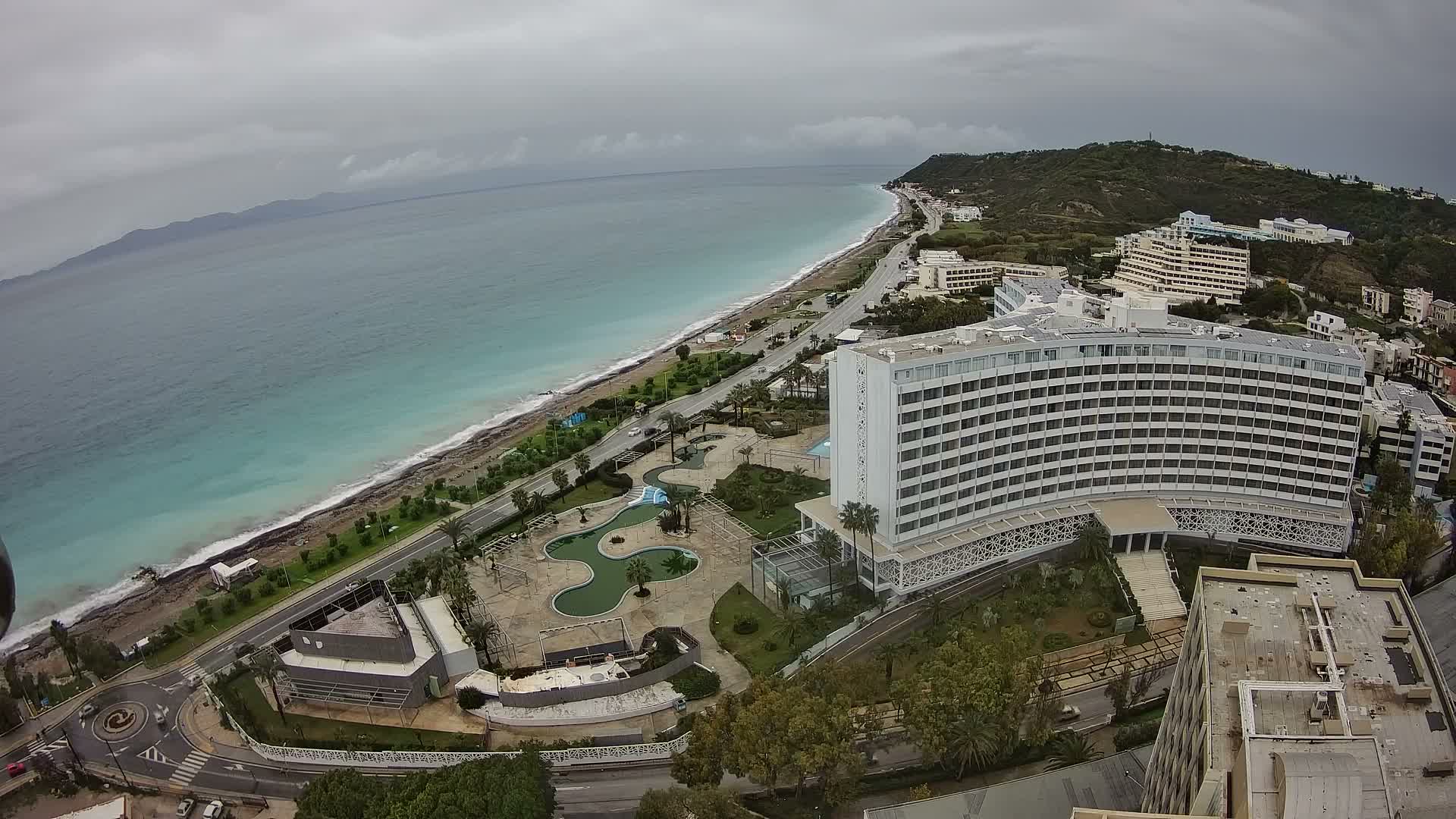 An overcast aerial view reveals a long stretch of coastline with turquoise waters, a road, several large hotels with pool areas, and green hills under a grey, cloudy sky.