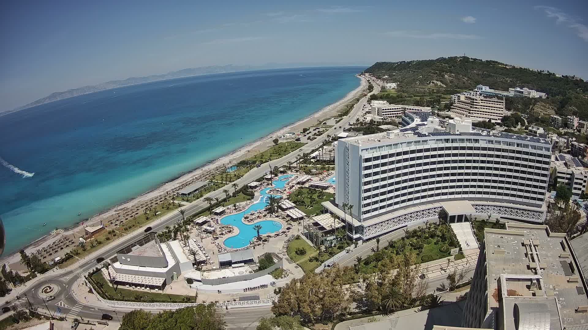 An overcast aerial view reveals a long stretch of coastline with turquoise waters, a road, several large hotels with pool areas, and green hills under a grey, cloudy sky.