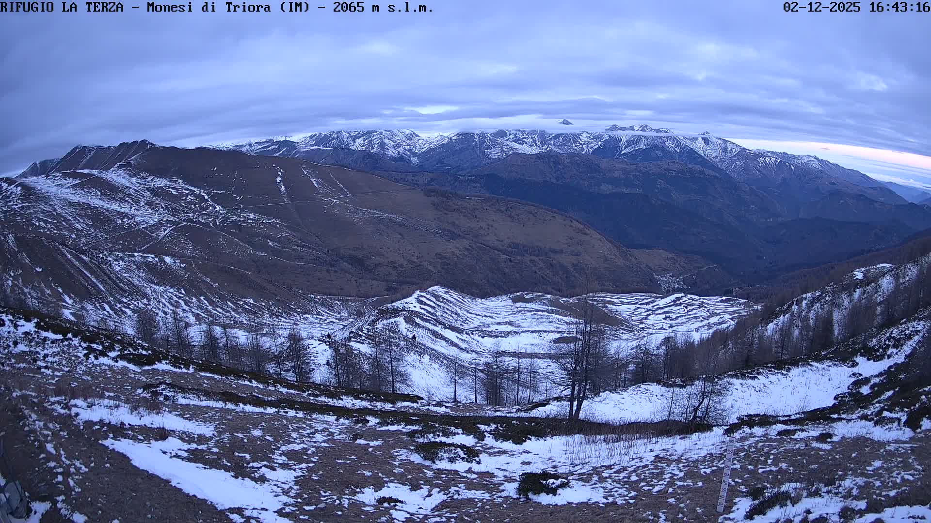 A panoramic view of a snow-dusted mountainous landscape unfolds under a cloudy, overcast sky, with patches of snow on the foreground slopes and snow-capped peaks in the distance.