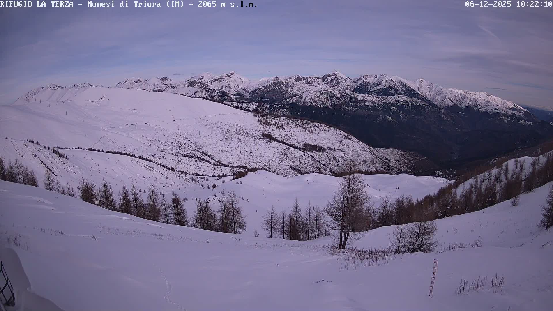 A snowy mountain vista extends under a partly cloudy sky, showcasing snow-dusted bare trees on the slopes and rugged, snow-capped peaks in the background.