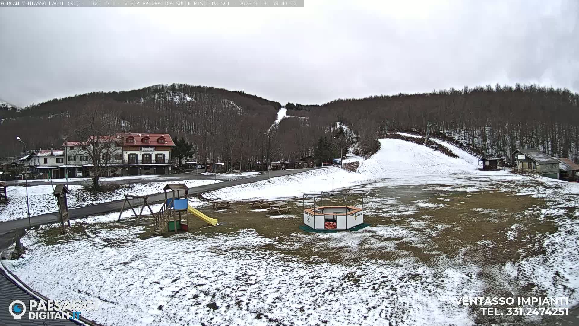 A snow-dusted playground and a building sit in a clearing near a hillside ski slope under an overcast sky.
