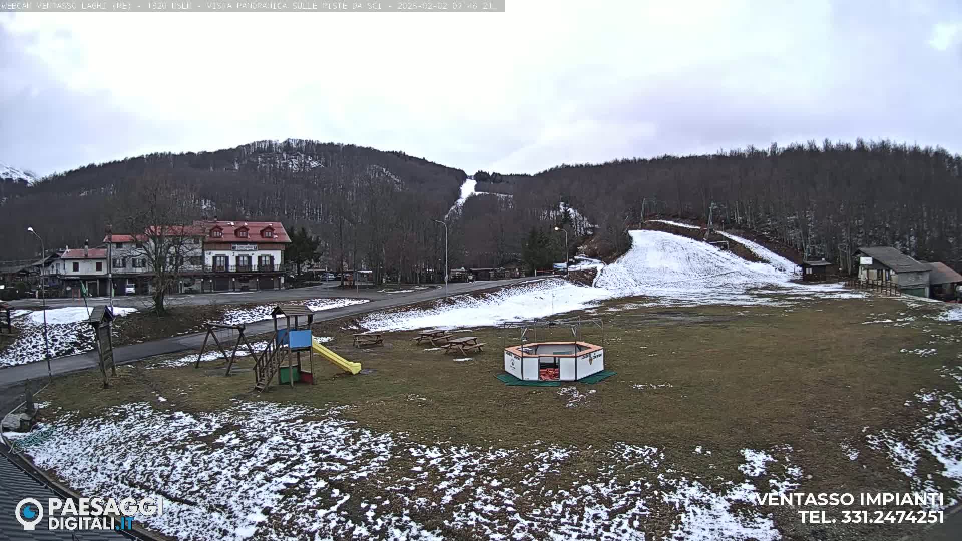 A snow-dusted playground and ski slope are situated below a wooded hillside under a cloudy sky.