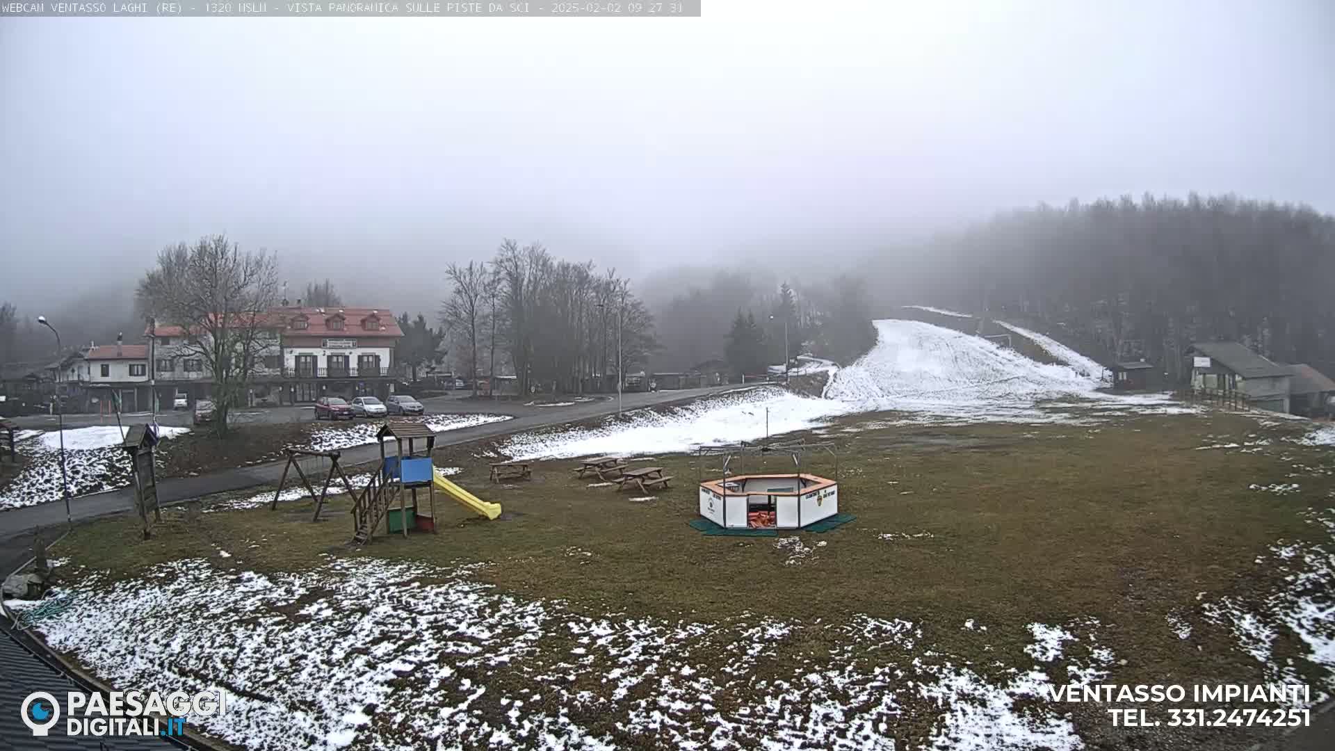 A foggy, snow-dusted landscape shows a small village with a building and playground near a ski slope.
