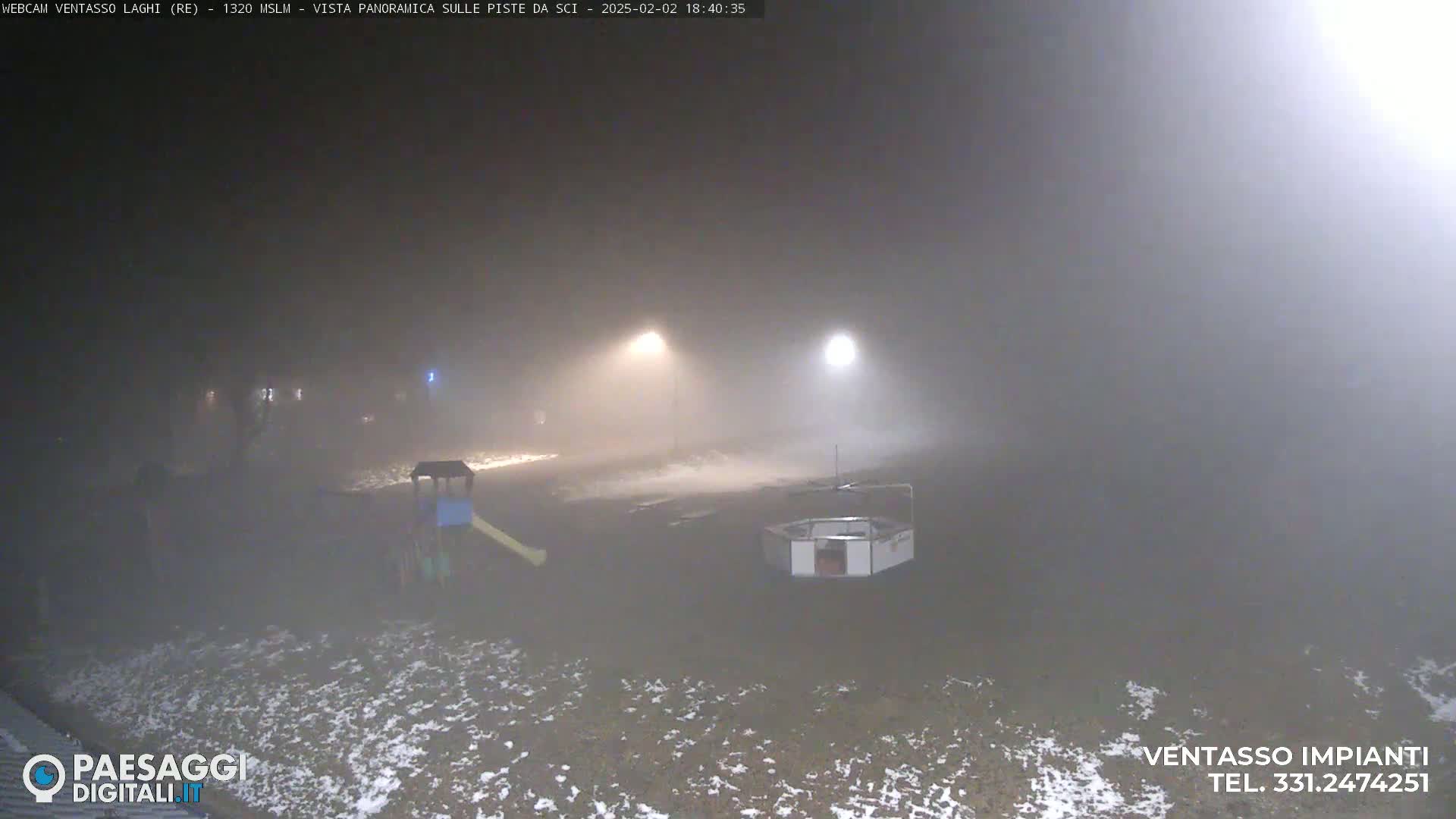 A foggy, nighttime scene shows a playground with a slide and a small structure in a snowy area illuminated by distant lights.