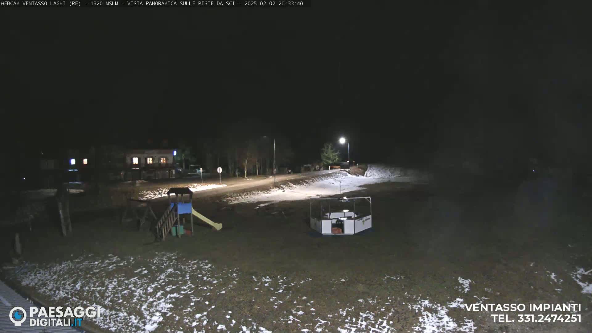 A nighttime view of a snow-dusted playground and a small building near a road, illuminated by streetlights.