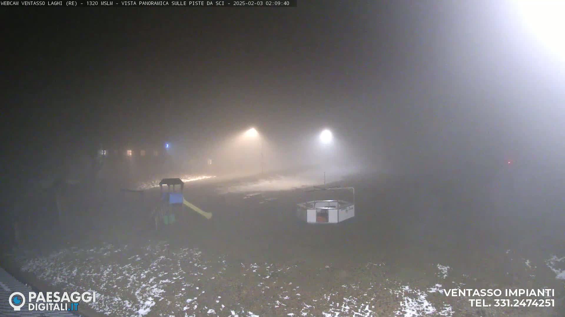 A foggy, snowy nighttime scene shows a playground with a slide and a small structure, illuminated by distant lights.