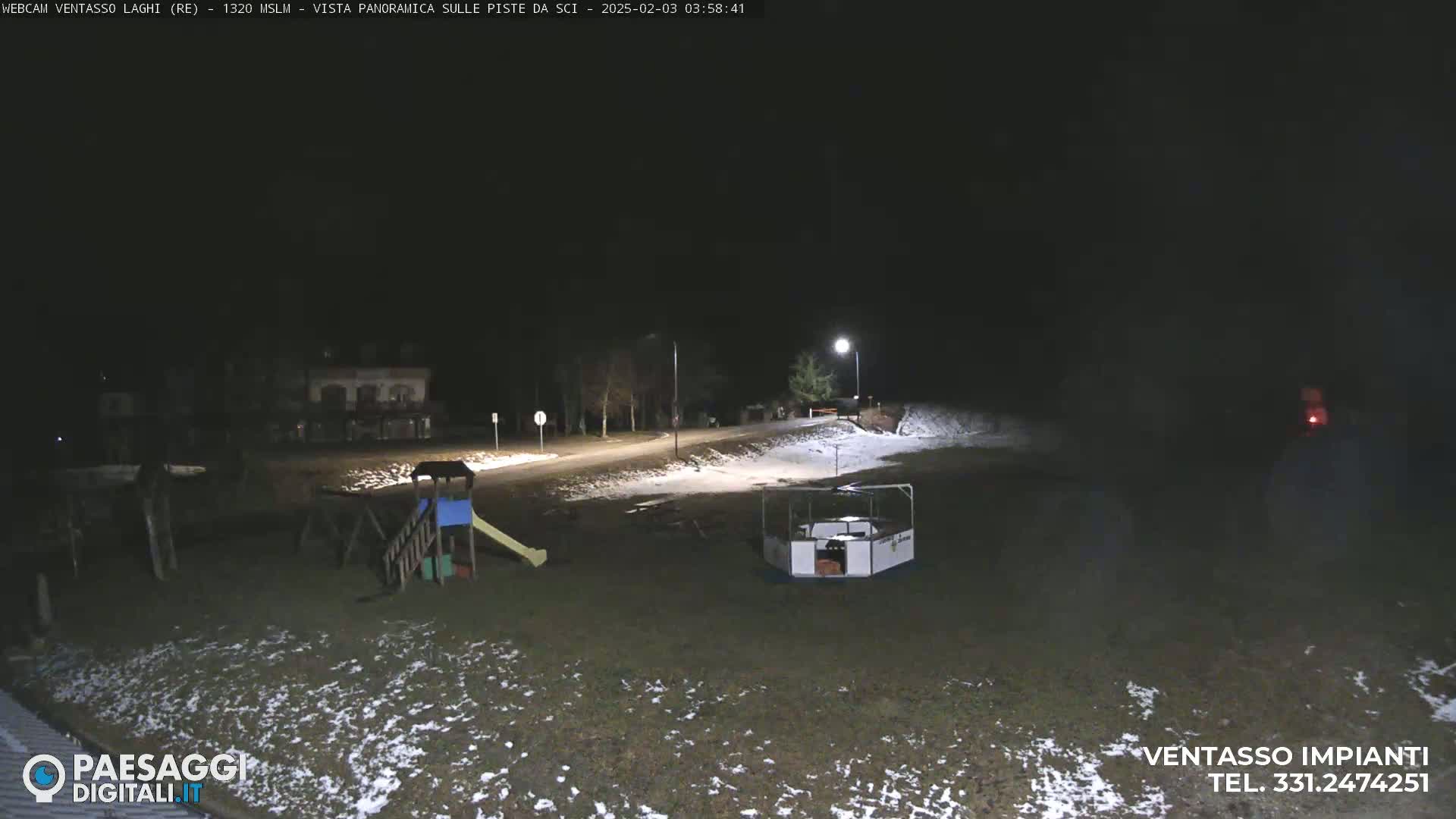 A nighttime view of a snow-dusted playground and empty road, illuminated by streetlights.