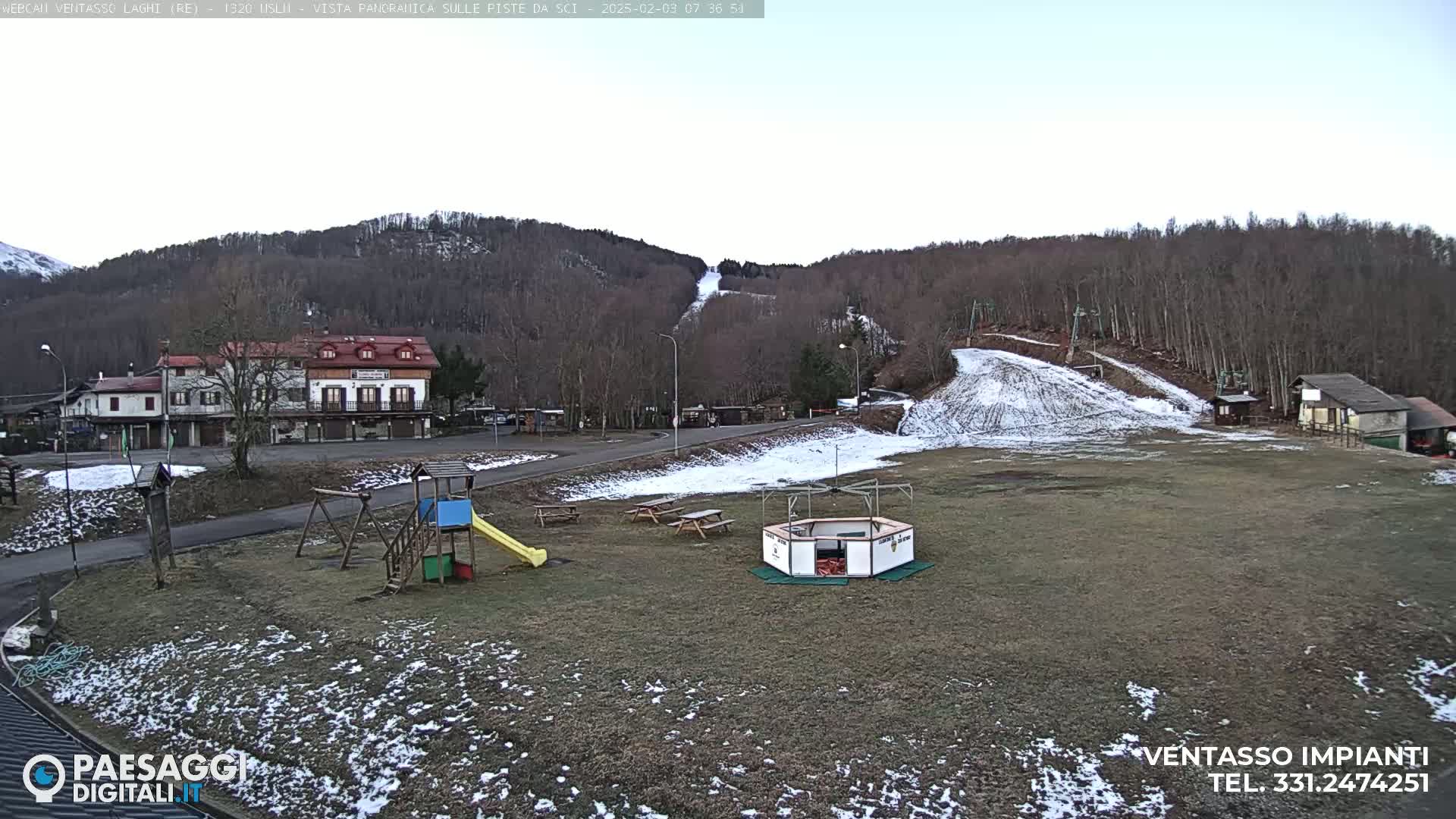 A mostly clear day reveals a snow-dusted ski slope in the background, a playground and picnic tables in the foreground, with a small village and buildings visible to the left.
