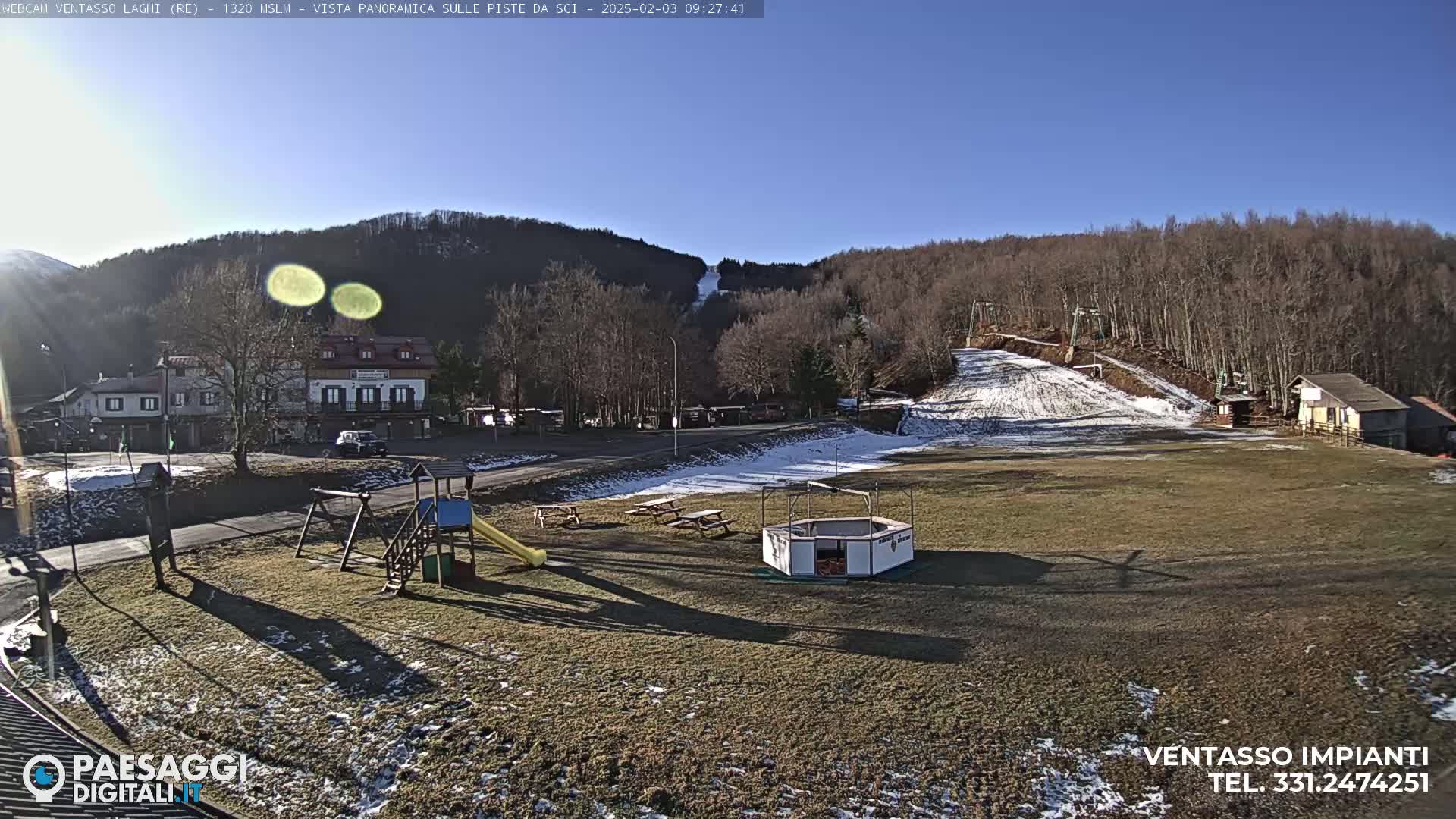 A sunny day reveals a ski slope with some snow, a playground, several buildings, and picnic tables in a grassy area, all nestled against a backdrop of leafless trees and hills.