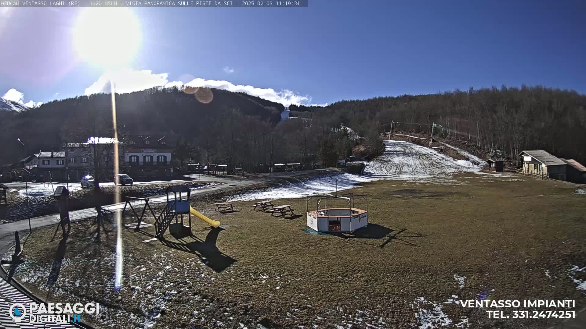 A sunny day reveals a playground, picnic tables, and a small building near a ski slope with some snow, set against a backdrop of trees and mountains under a mostly clear sky.