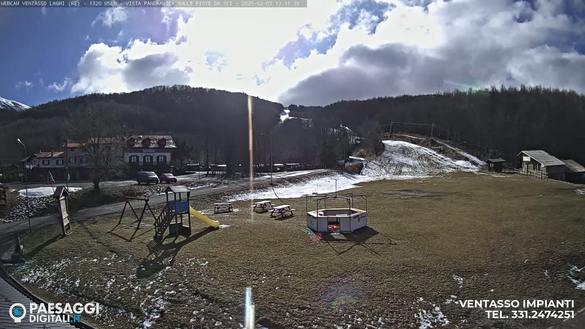 A partially sunny day shows a playground, picnic tables, and a small building in a grassy area near a ski slope and a cluster of buildings, all set against a backdrop of a wooded mountain.