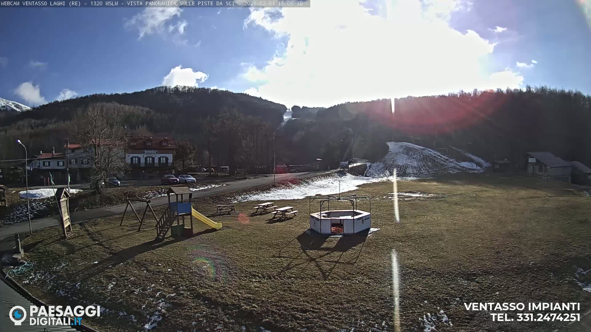 A sunny, partially cloudy day reveals a grassy area with a playground, picnic tables, and a small structure near buildings and a ski slope in the background.