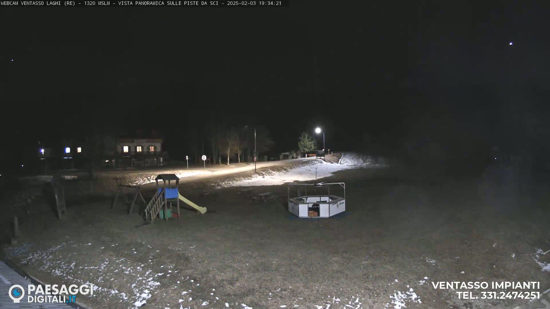 A nighttime view of a playground and a small structure in a snow-dusted area, illuminated by artificial lights under a clear, dark sky.