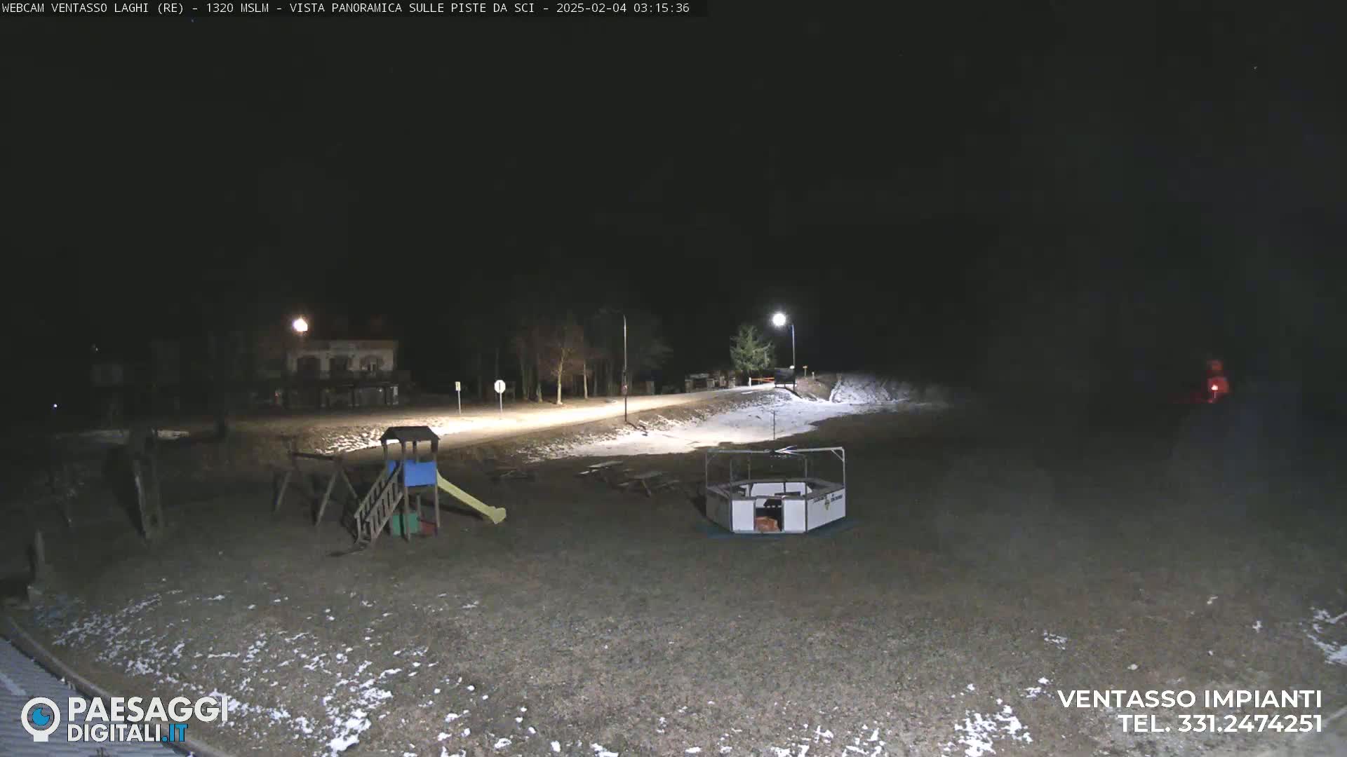A nighttime view of a playground and a small roundabout in a snow-dusted area, illuminated by streetlights.
