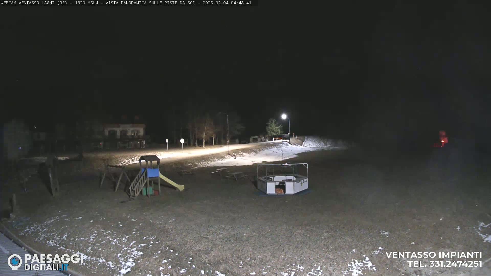 A nighttime view of a snow-dusted playground and a paved area, lit by lampposts, with some trees in the background.
