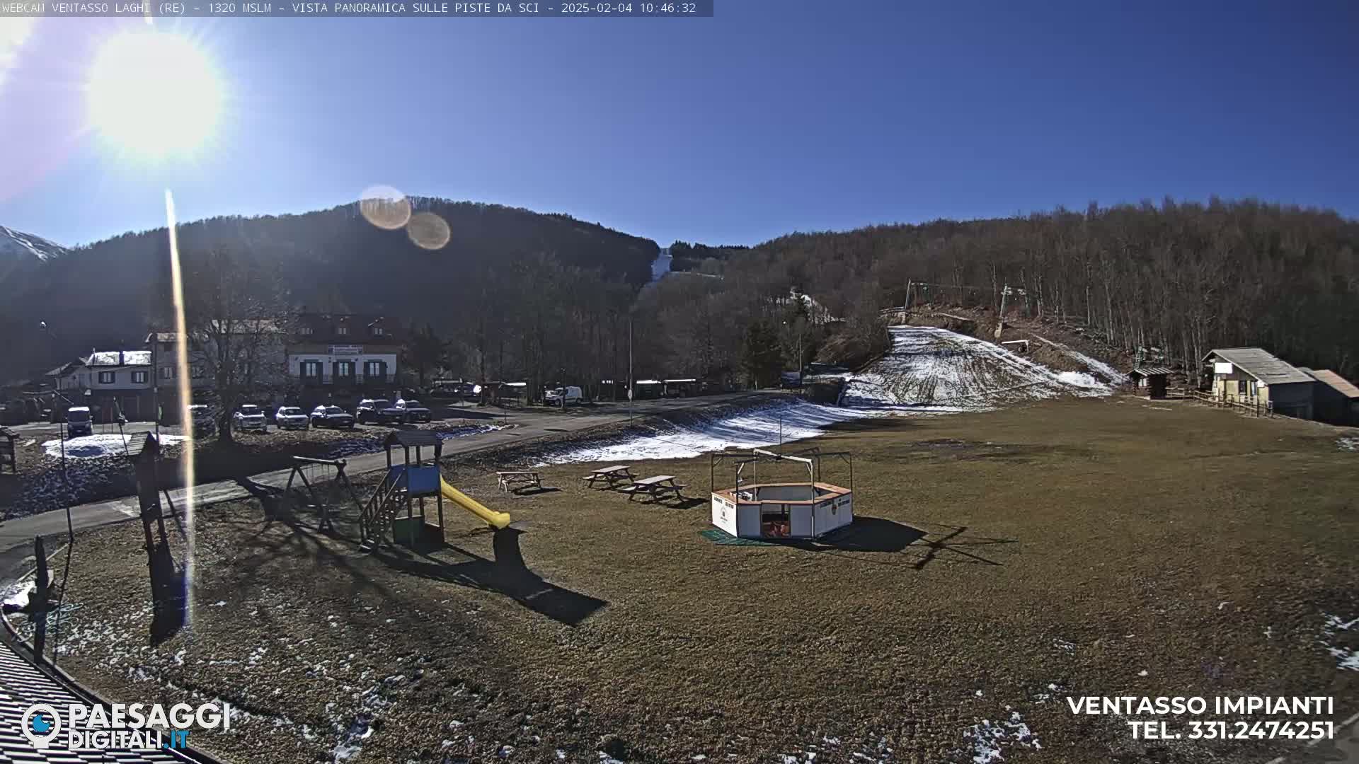 A sunny day at a ski resort shows a playground, picnic tables, and a small building near a grassy area with a ski slope and several buildings in the background.