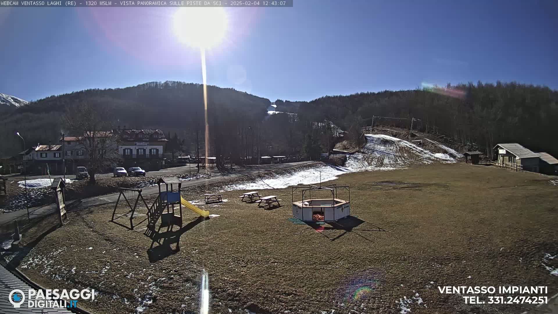 A sunny day at a ski resort shows a playground, picnic tables, a small building, and a ski slope in the background.
