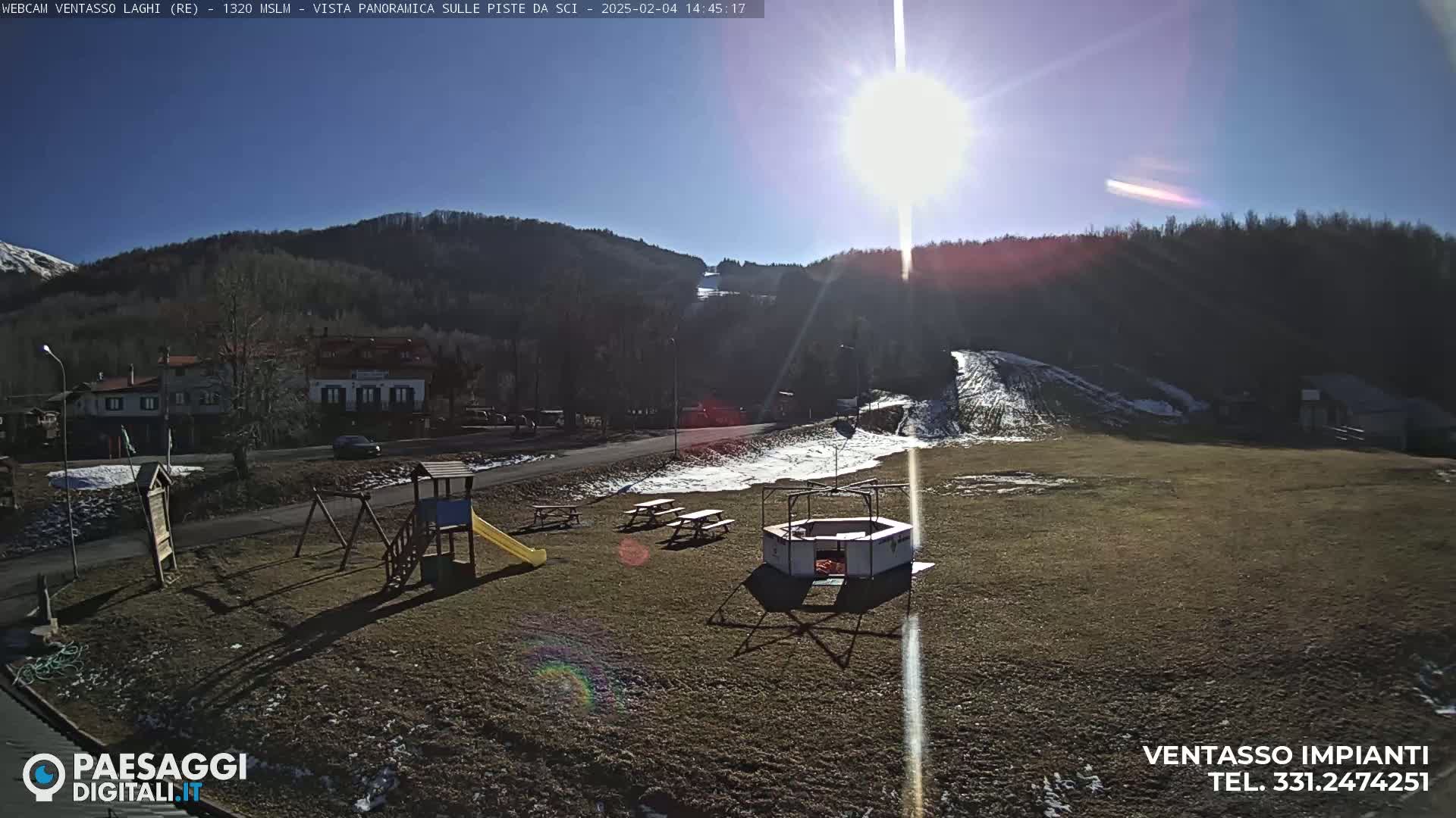A sunny day reveals a playground, picnic tables, and a small structure in a grassy area near a road and a hillside with some snow and trees.