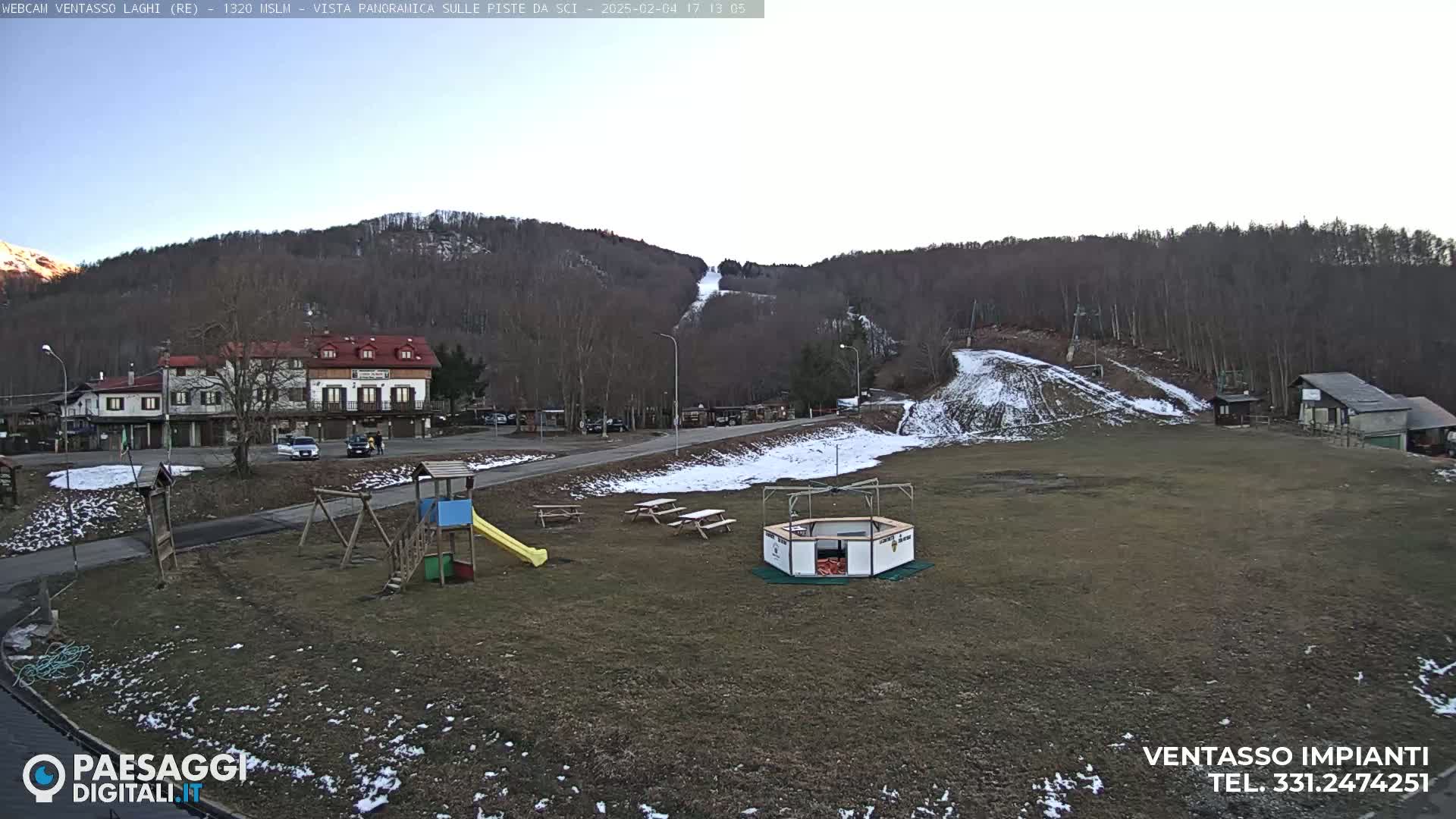 A mostly clear, winter day reveals a snow-dusted ski slope behind a small village with buildings, a playground, and picnic tables.