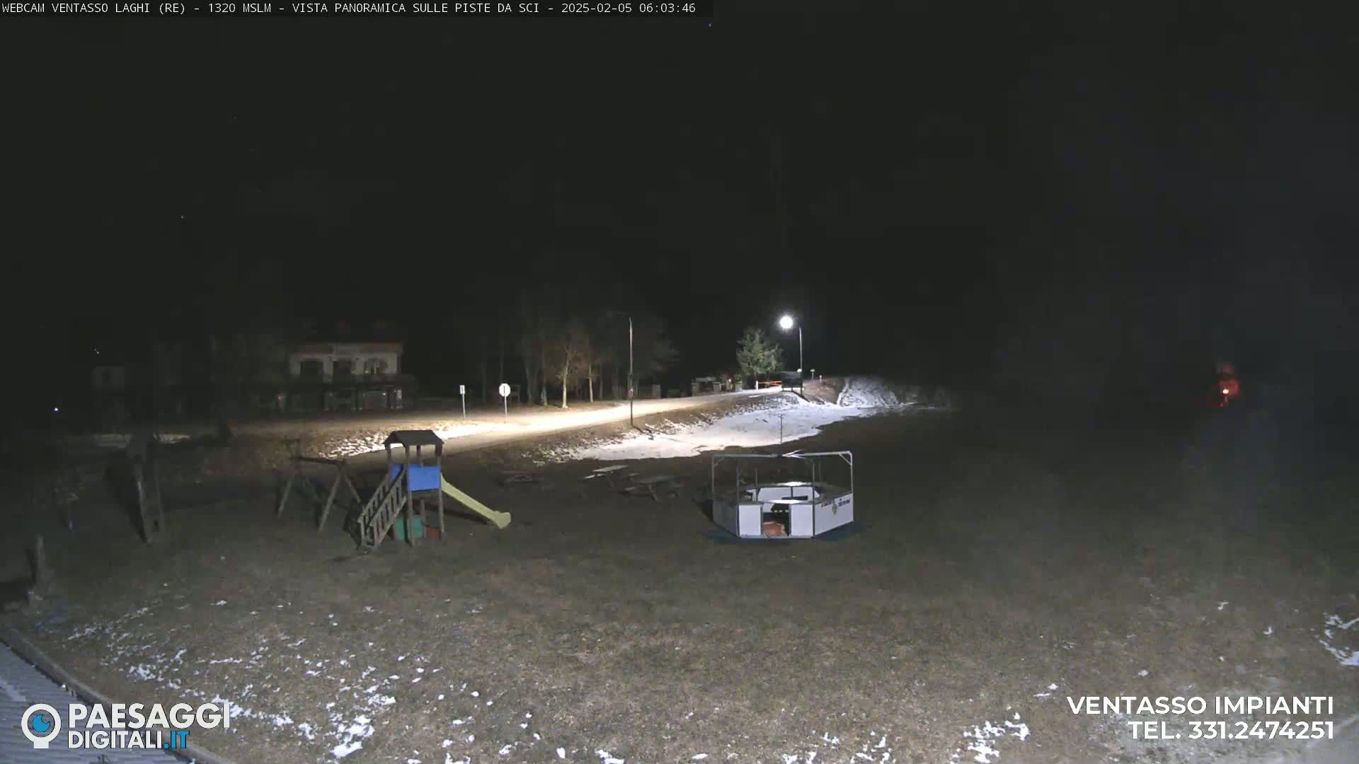 A nighttime view of a playground and a small structure, with a road and some sparsely-treed areas in the background, all covered in a light dusting of snow.