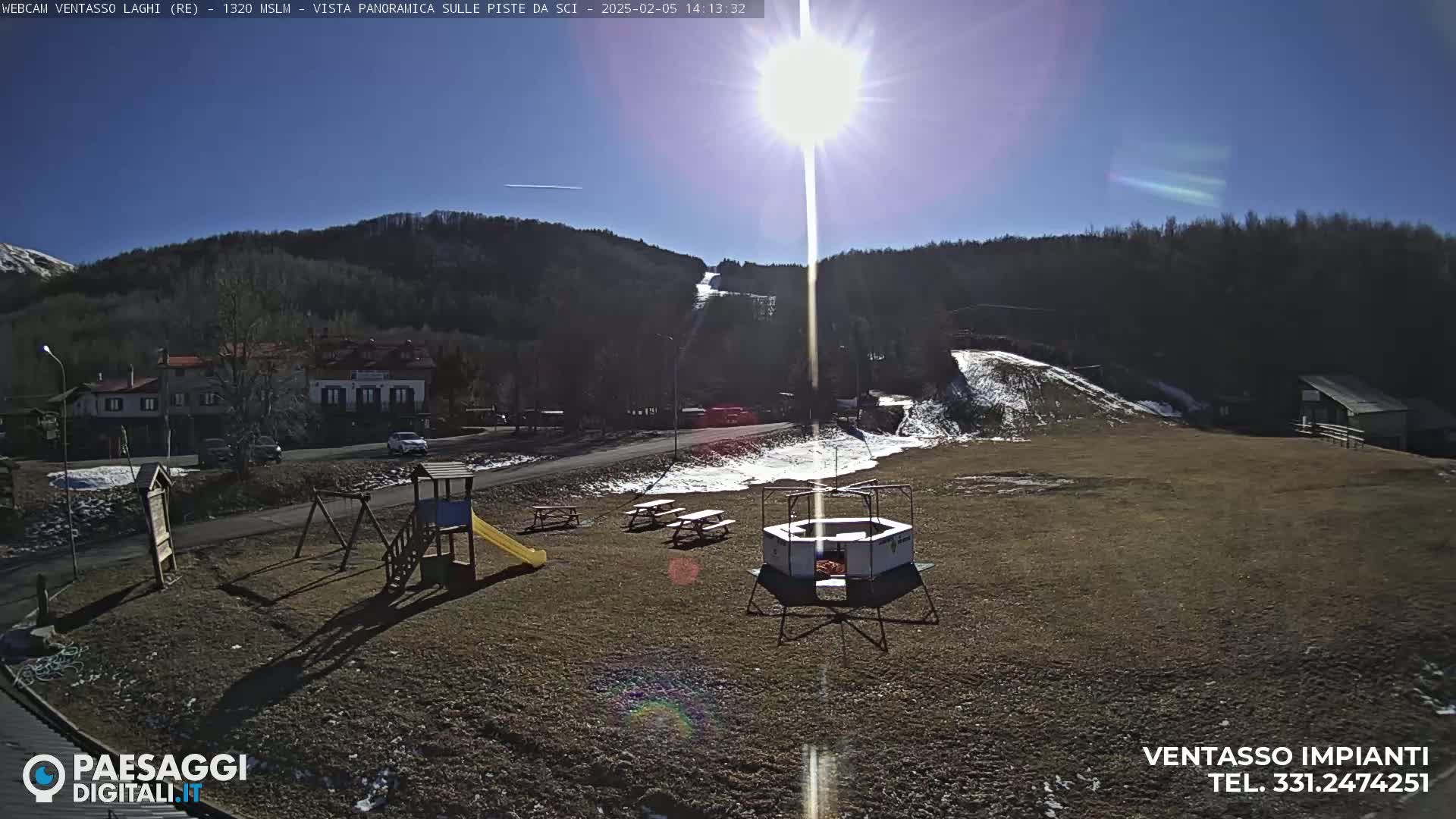 A sunny day reveals a playground, picnic tables, and a ski slope with some snow, all situated in front of a hillside covered in bare trees and a small village.