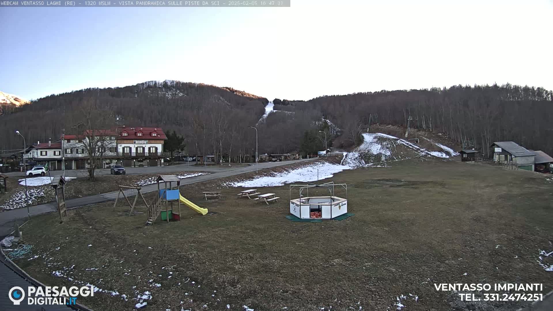 A mostly sunny view of a ski resort with a playground, picnic tables, and a small building, with patches of snow on the ground and a ski slope visible in the background.