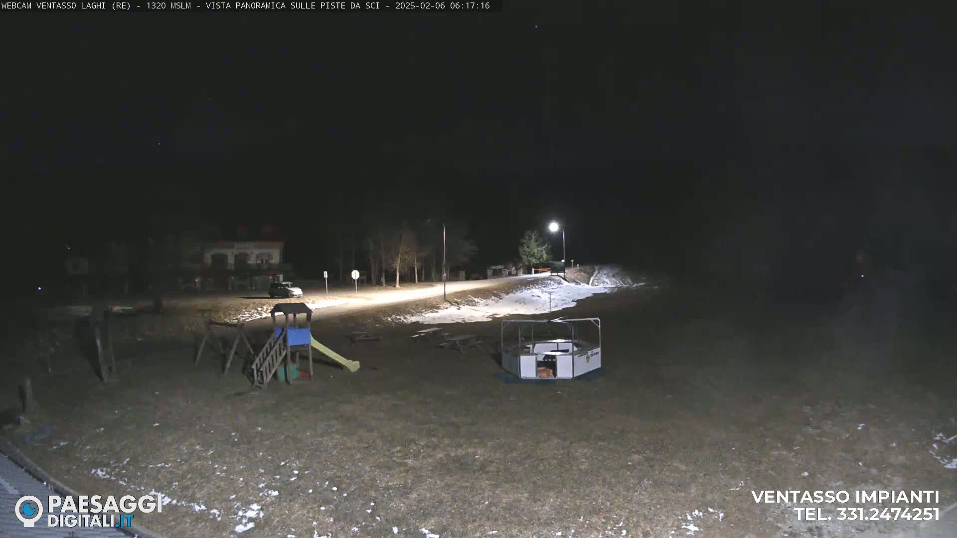 A nighttime view of a playground and a small structure, with patches of snow on the ground under a dark sky.
