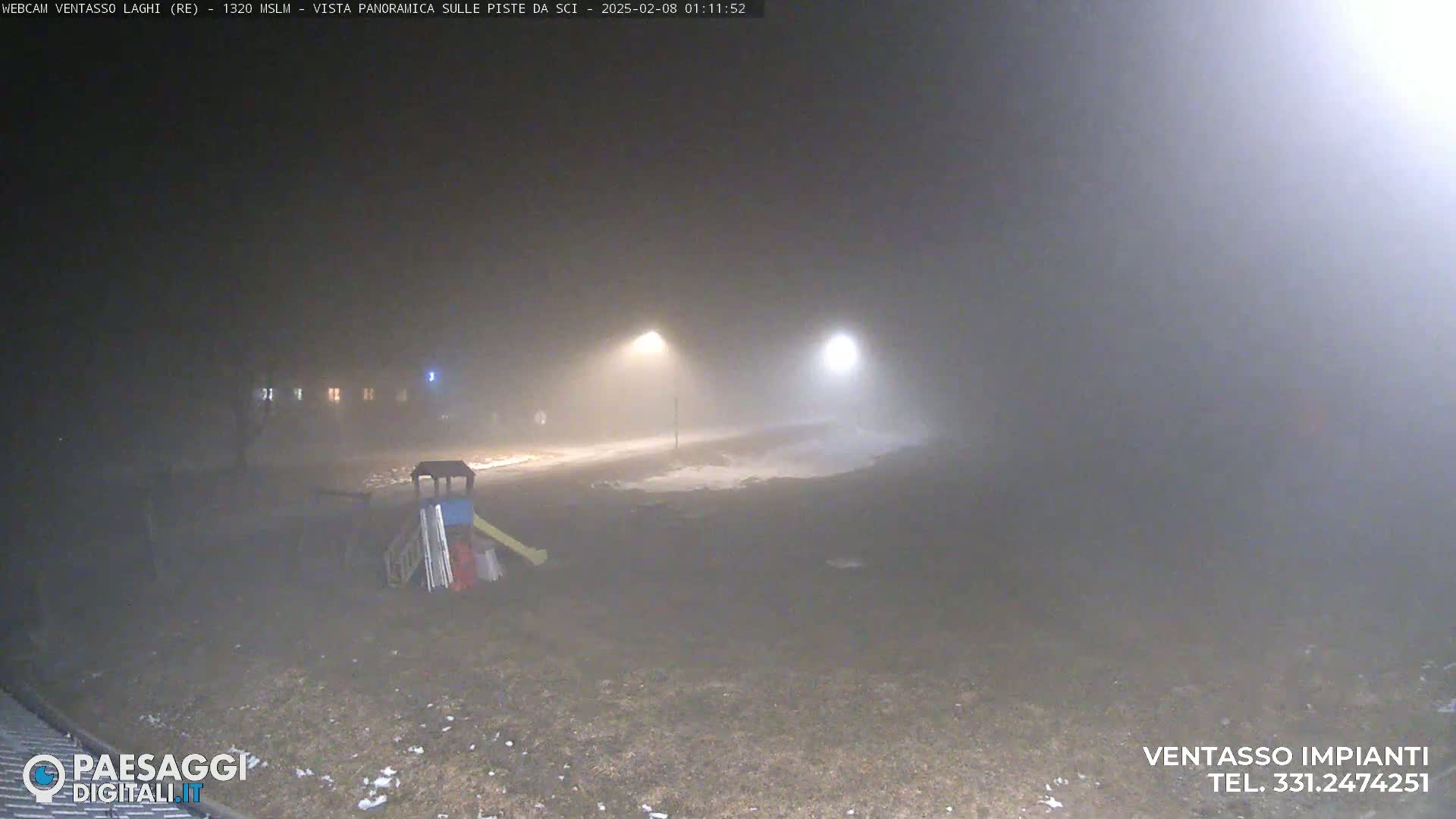 A foggy nighttime scene shows a playground with a slide, sparsely lit by streetlights, and a small amount of snow on the ground.