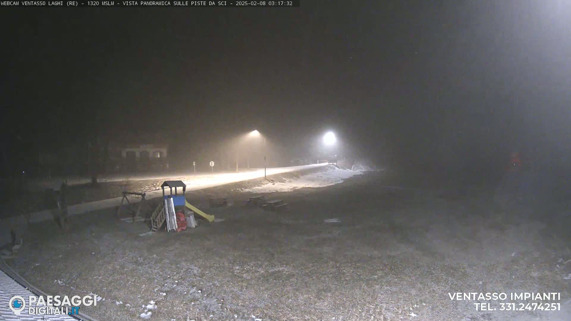 A nighttime, snowy scene shows a playground and a paved path lit by streetlights.