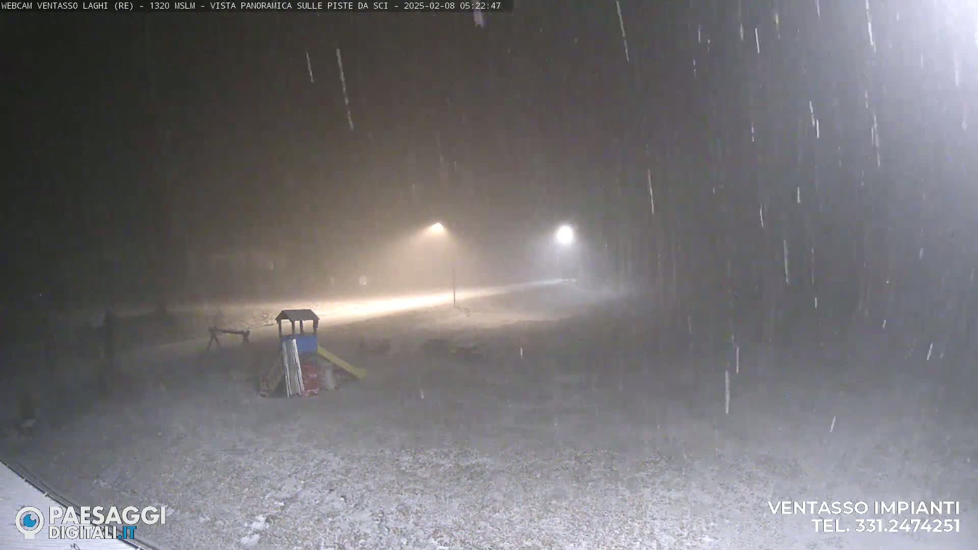 A snow-covered playground is dimly lit by two streetlights under heavy snowfall at night.