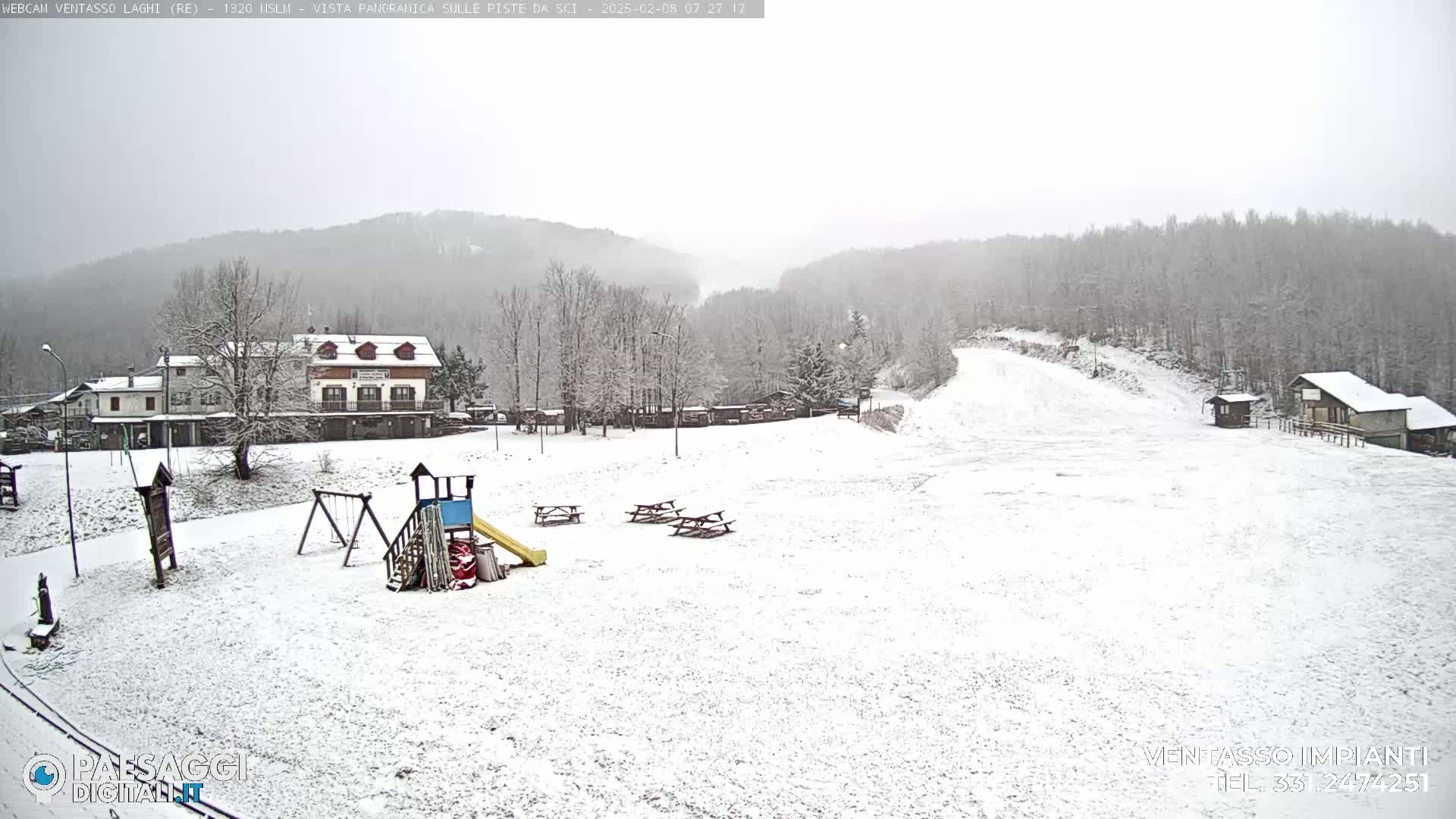A snow-covered playground and ski slope are visible beneath a lightly falling snow, with snow-covered buildings and trees in the background.