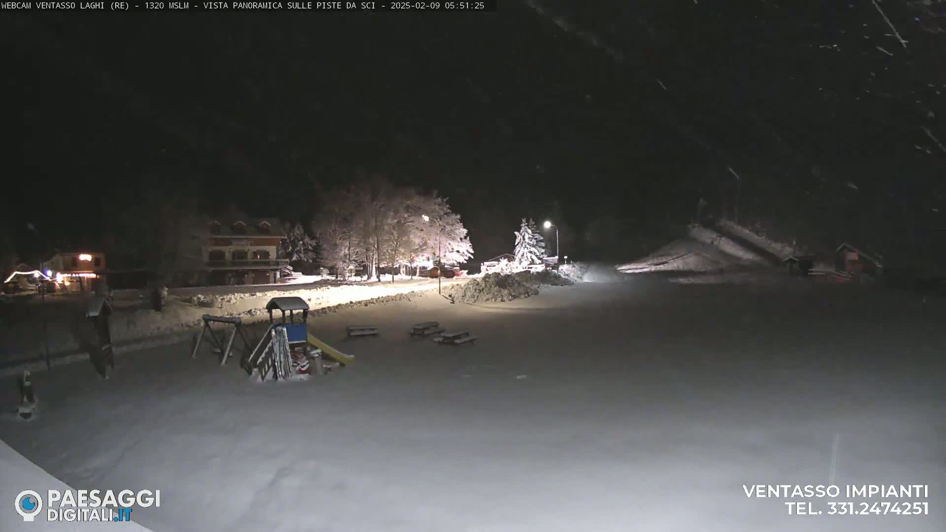 A snow-covered playground and picnic area are visible at night under falling snow near some buildings and trees.