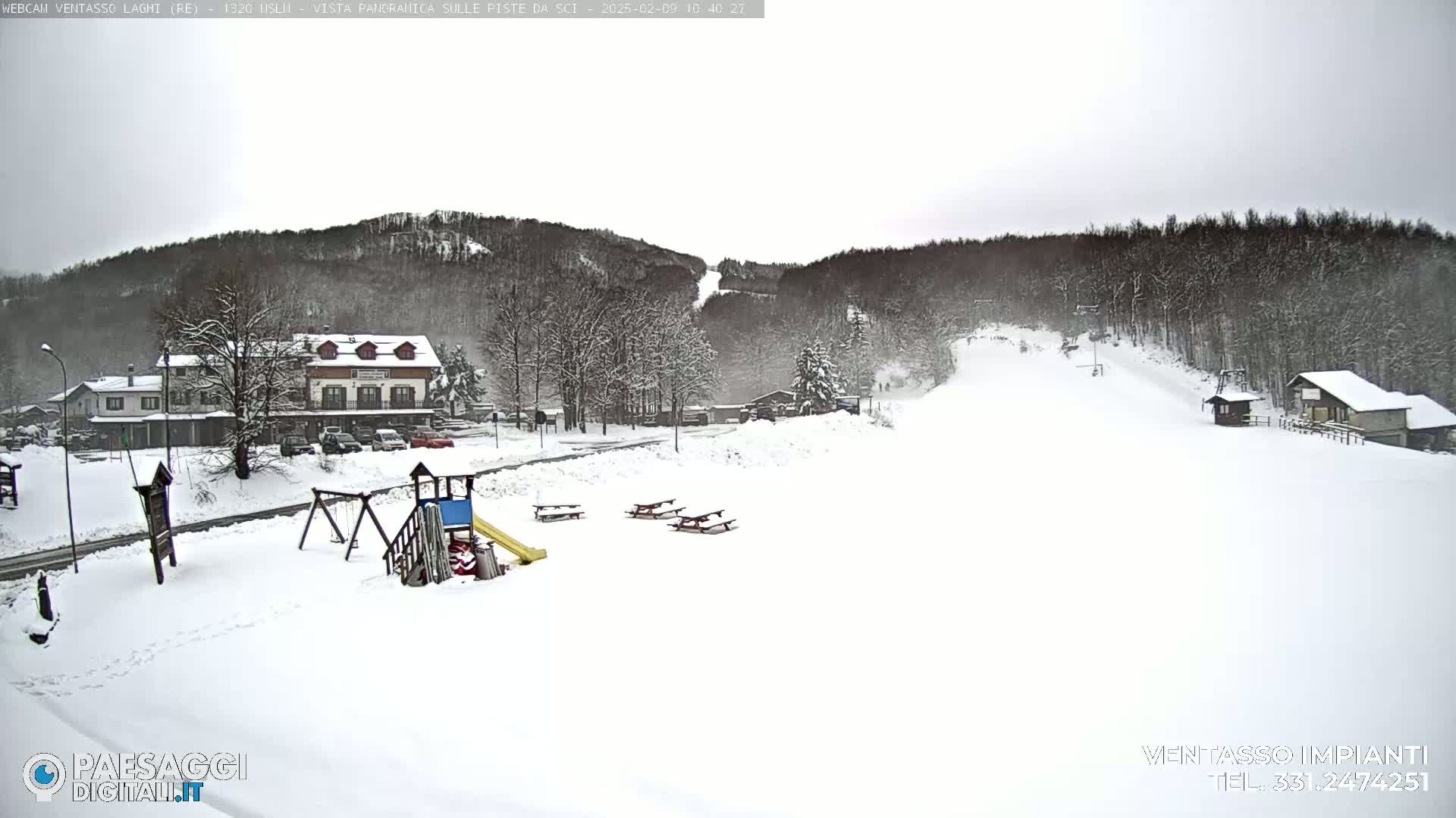 A snow-covered landscape features a small village with buildings, a playground, picnic tables, and a ski slope on a cloudy day.