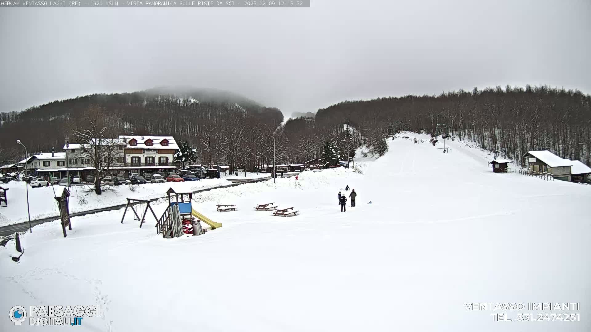 A snow-covered landscape features a ski slope, a few people, picnic tables, a playground, buildings, and a wooded hillside under an overcast sky.