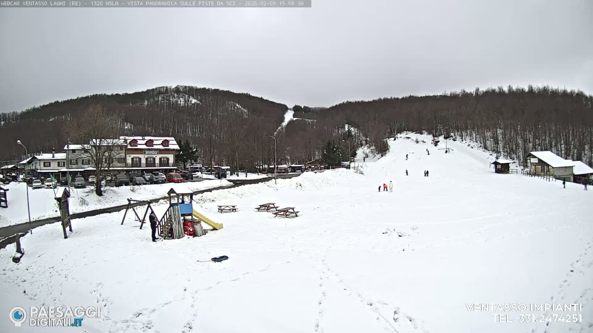 A snow-covered ski slope with several people skiing and snowboarding, a playground, picnic tables, and buildings in the background under an overcast sky.