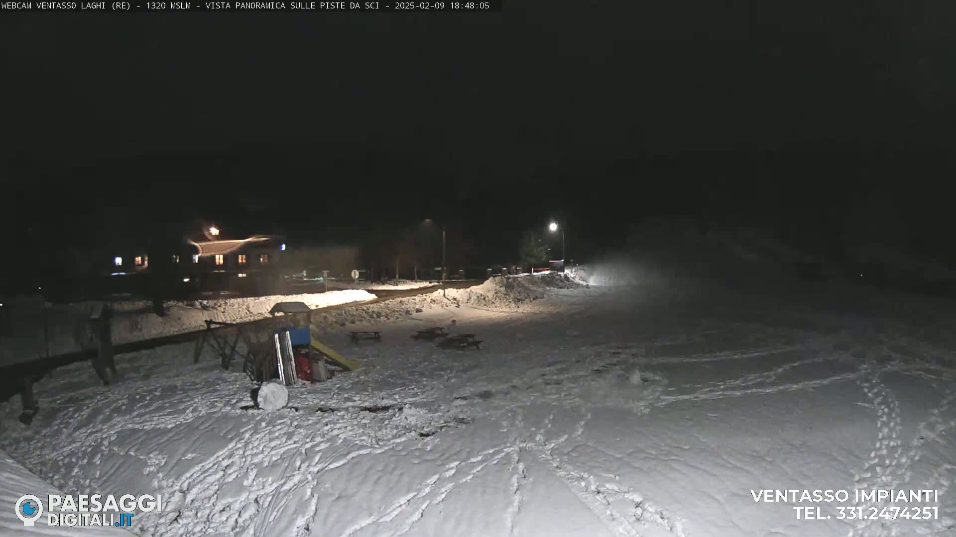 A snow-covered area at night is illuminated by artificial lights, showing a building in the distance and playground equipment in the foreground.