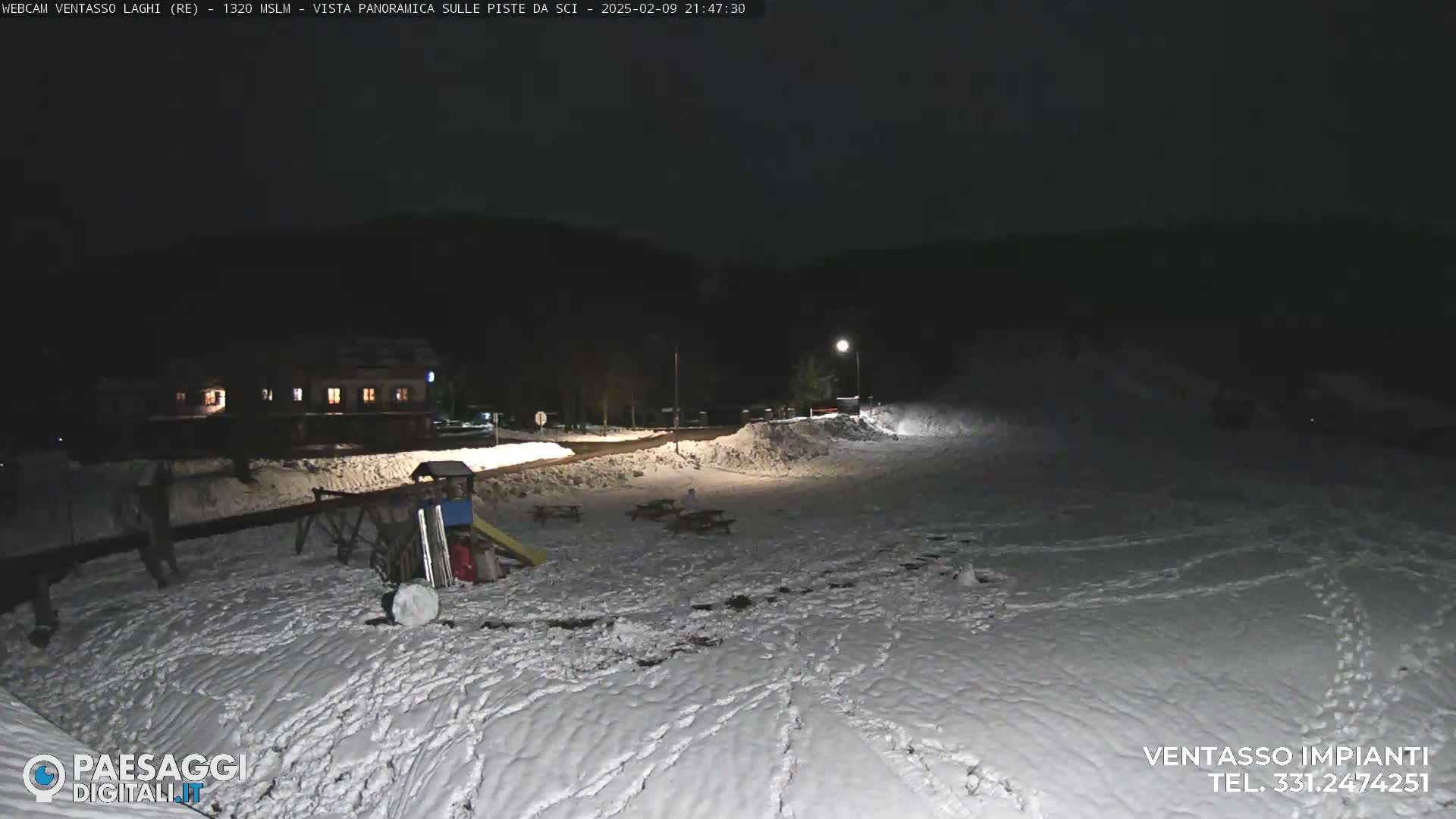 A snow-covered area at night, illuminated by a single lamppost, shows a playground and a building in the distance.