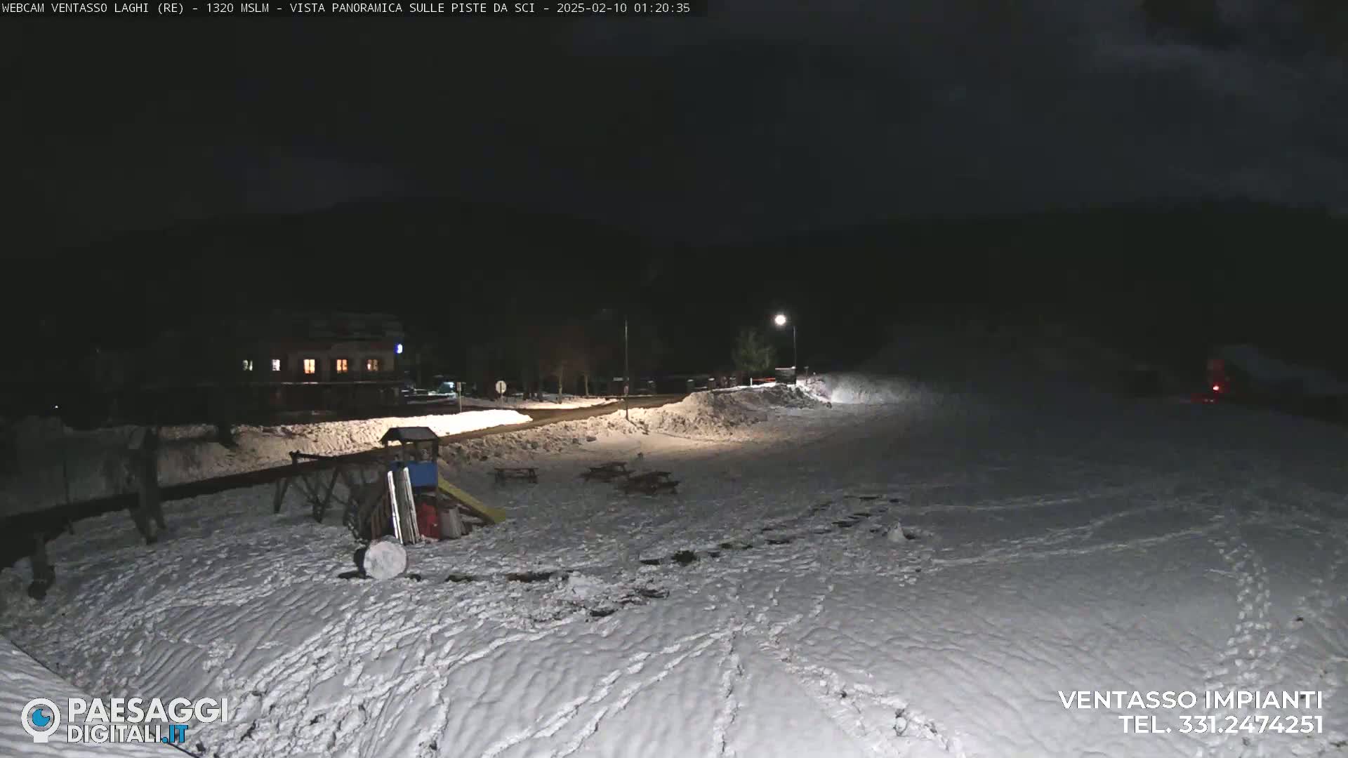 A snow-covered area at night, illuminated by a lamp, shows a building in the distance, some playground equipment, and footprints in the snow.