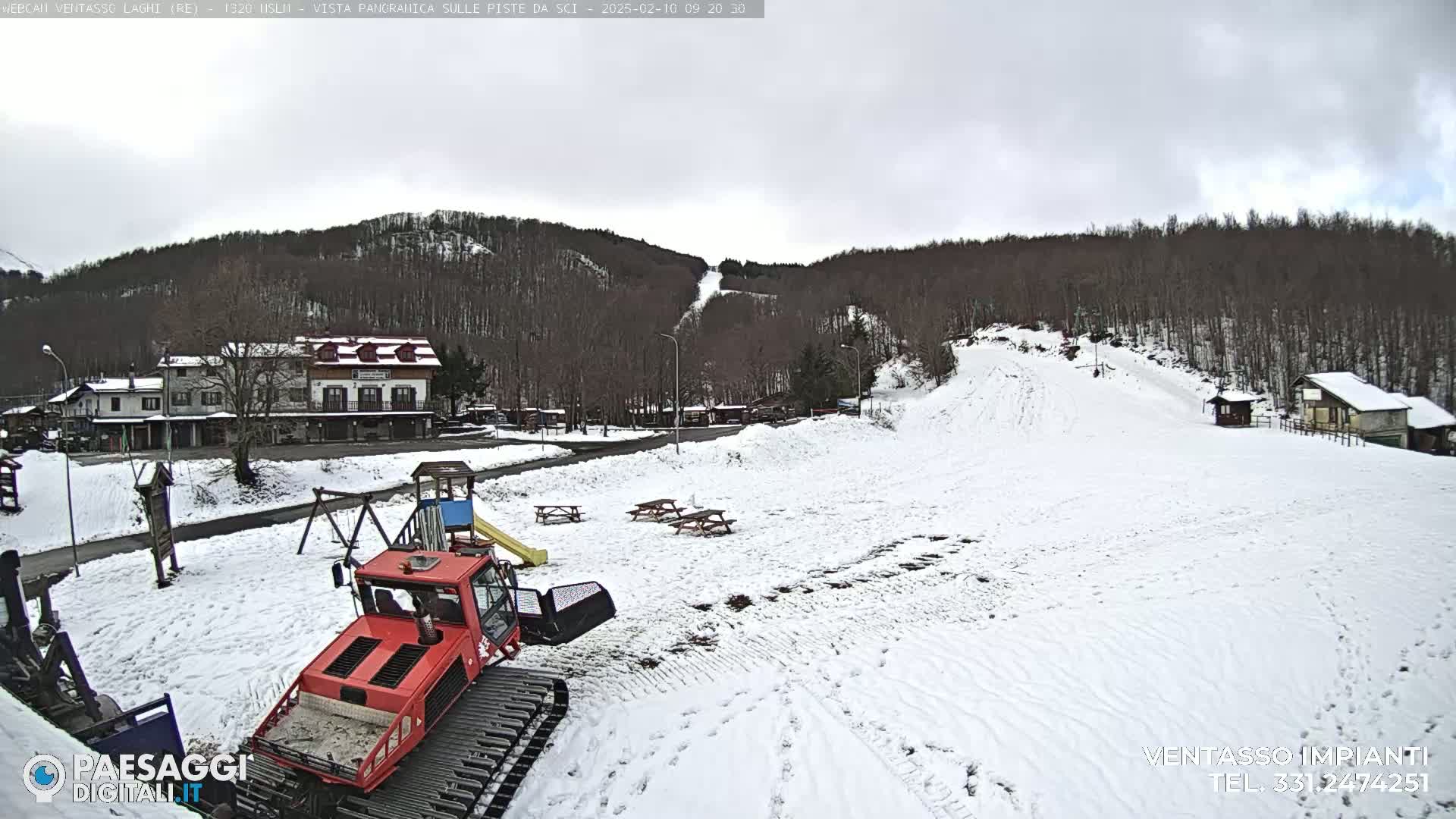 A snow-covered landscape with a red snowcat, playground equipment, picnic tables, and buildings, all set against a backdrop of a wooded hillside under an overcast sky.
