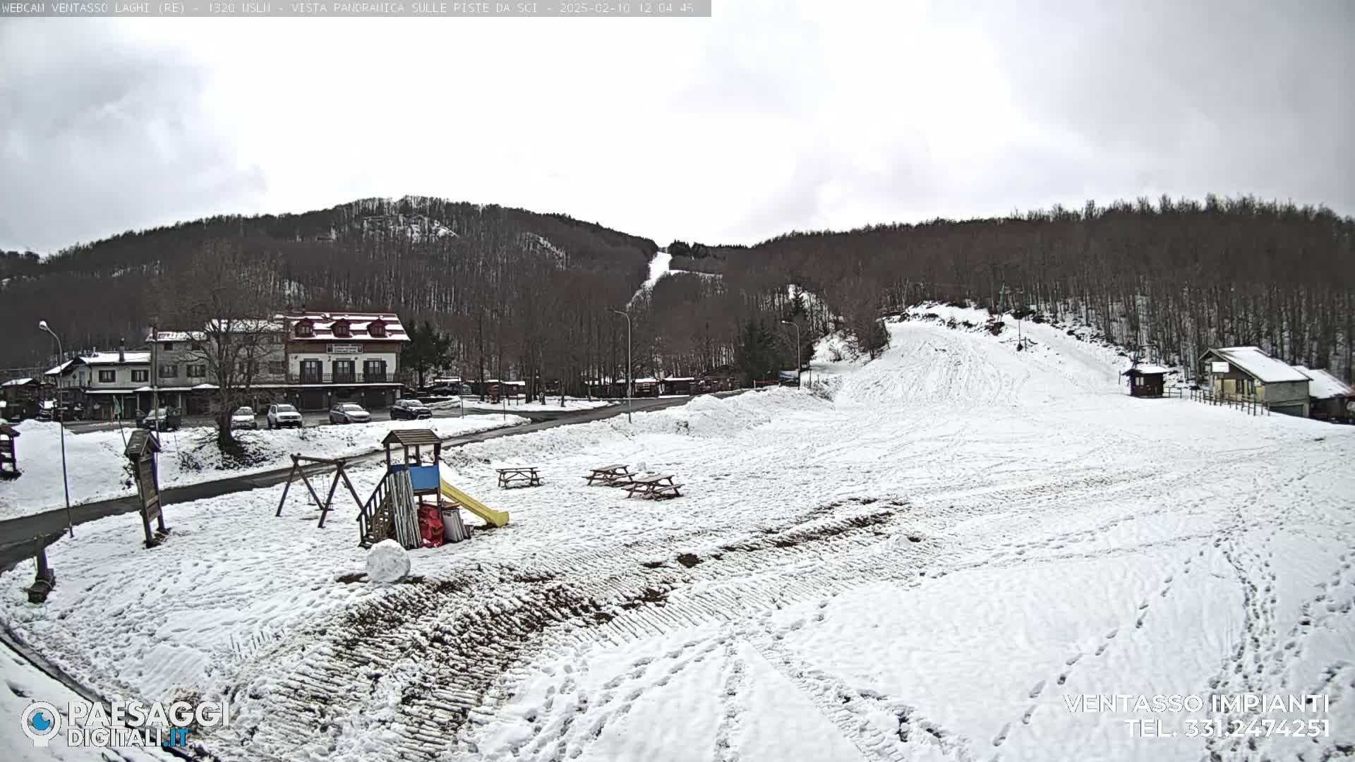 A snow-covered playground and picnic area sit below a snow-dusted ski slope and several buildings, all nestled against a backdrop of bare, snow-capped trees under an overcast sky.