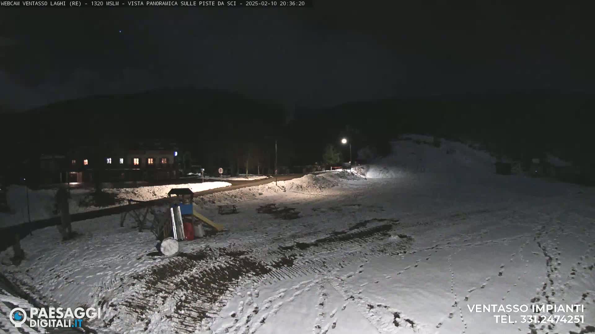 A snow-covered area at night, illuminated by lights, shows a building in the background and playground equipment in the foreground.