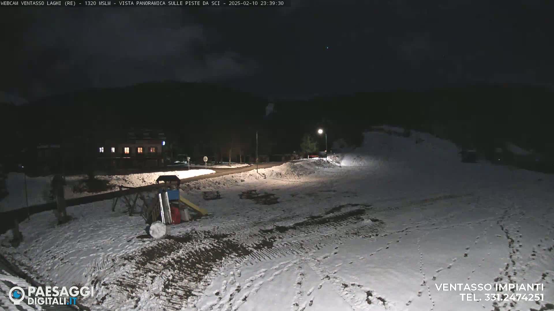 A snow-covered area at night, illuminated by a streetlight, shows a small building, playground equipment, and a road.