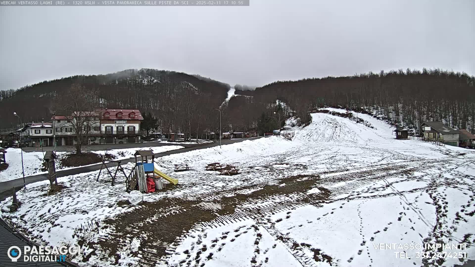 A snow-covered ski slope with a few buildings and a playground in the foreground, under an overcast sky.