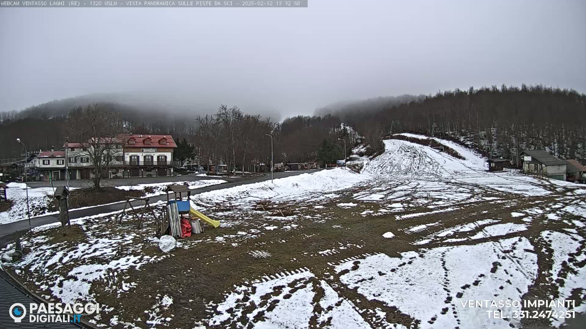 A partially snow-covered ski resort area with a building, playground equipment, and a ski slope under a foggy sky.